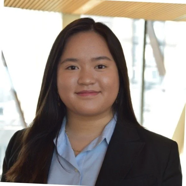 A young woman with long dark hair wearing a black blazer and light blue collared shirt, standing indoors with a blurred background of windows and wooden ceiling.