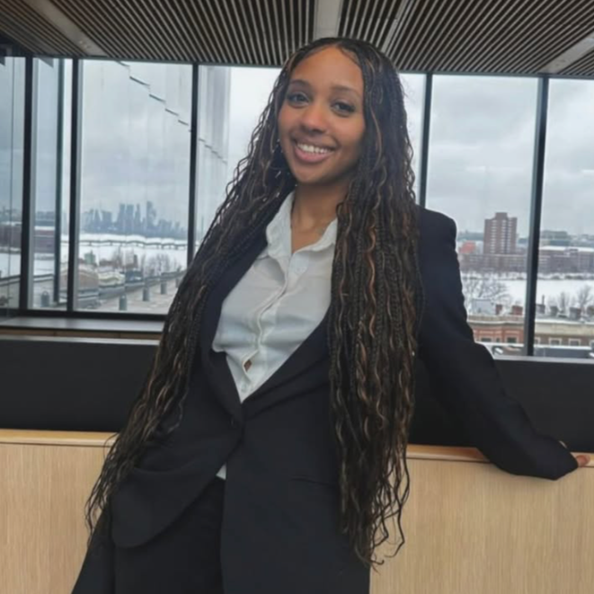 A young woman in business attire smiling, standing indoors with large windows showing a snowy cityscape and water in the background.