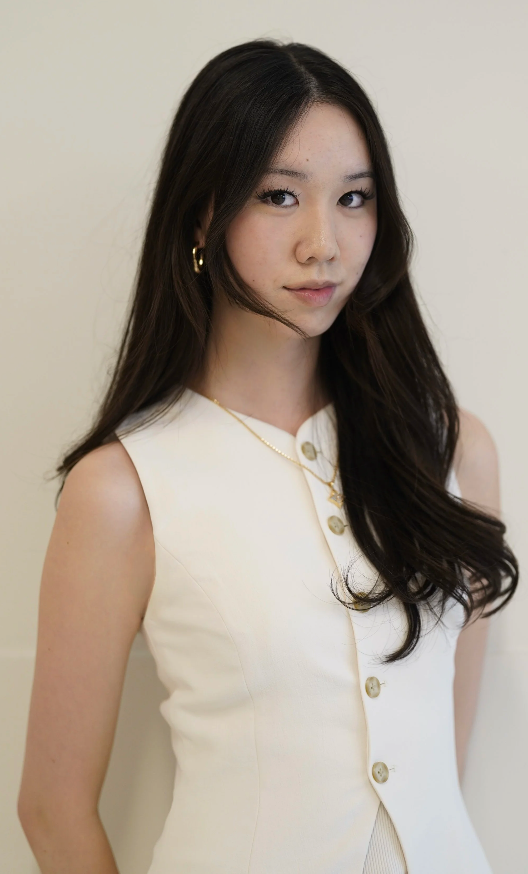 A woman with long dark hair wearing gold earrings and layered necklaces, dressed in a sleeveless white button-up outfit, standing against a plain off-white background.