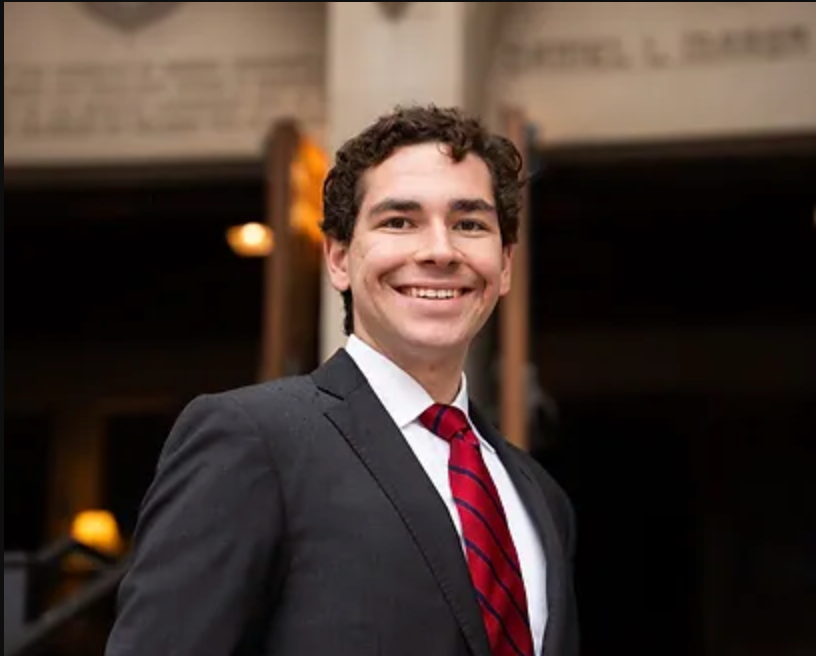 A smiling man in a dark suit, white shirt, and red striped tie stands outdoors at night, with a blurred background of lights and structures.