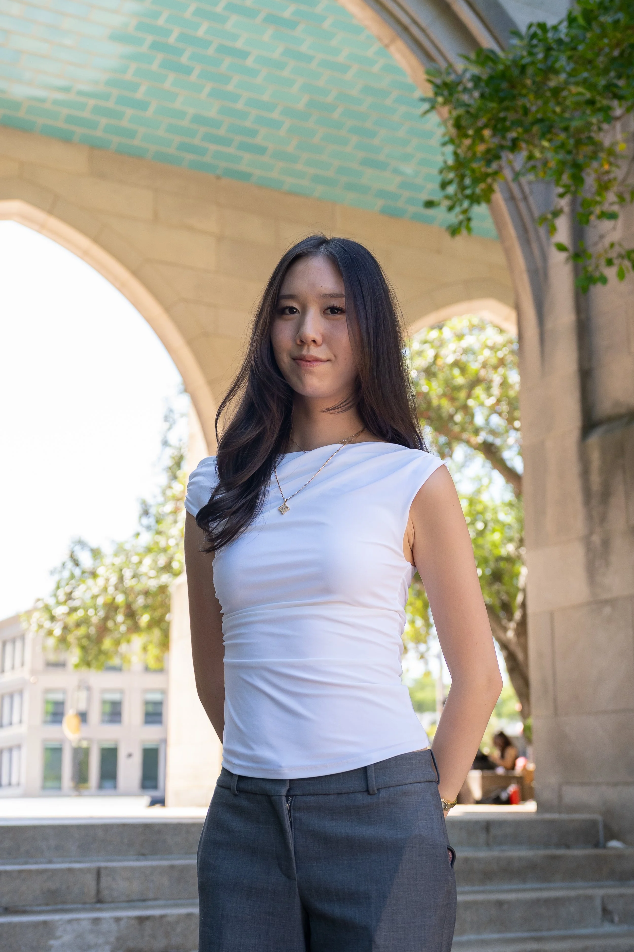 A young woman with long wavy dark hair, wearing a sleeveless white top with buttons and gold jewelry, standing against a plain light-colored background.