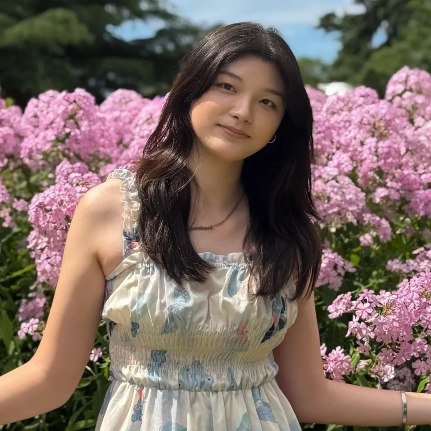 A young woman with long dark hair, wearing a sleeveless light dress, standing in front of pink hydrangea flowers outdoors on a sunny day.