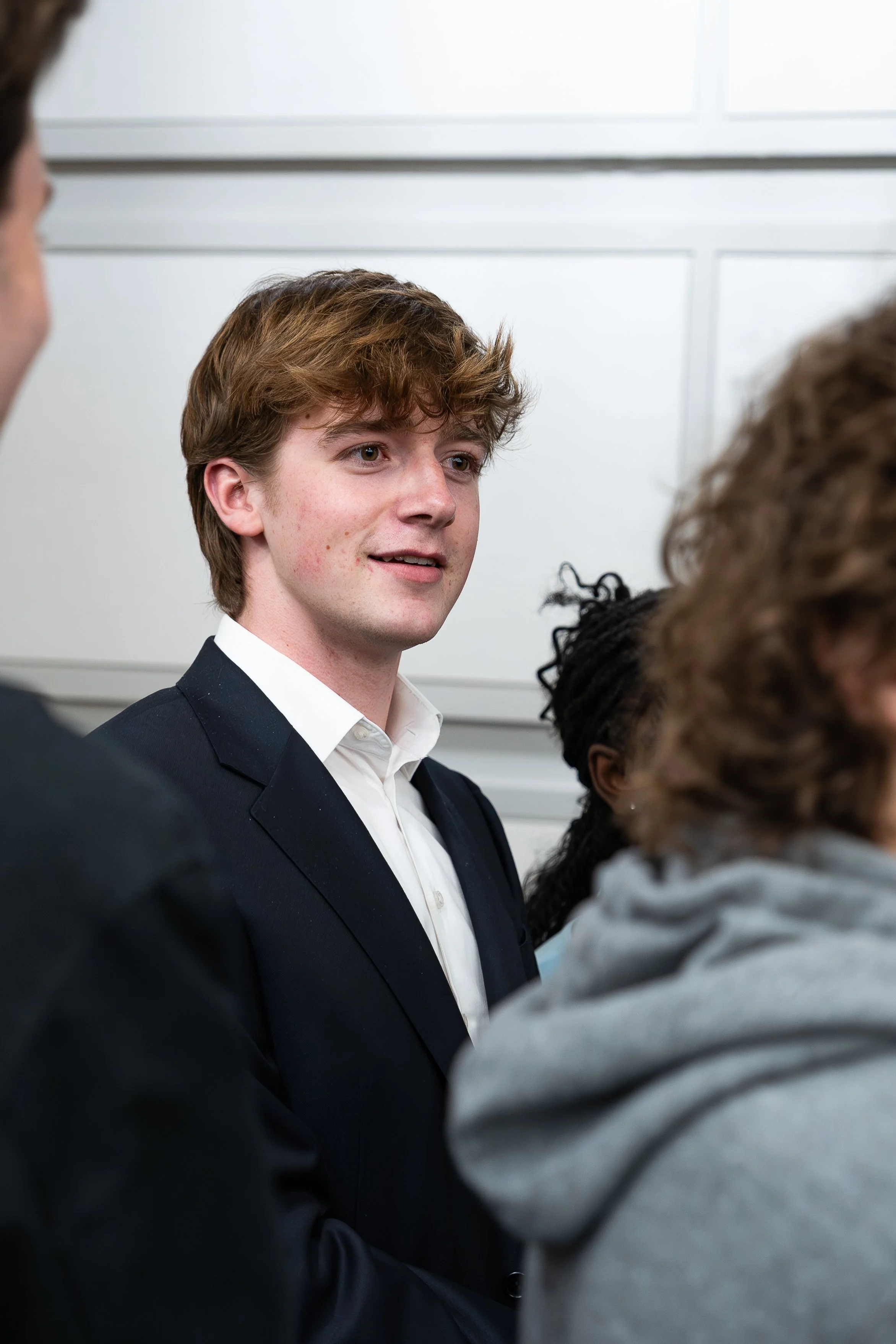 A young man in a black suit and white shirt engaged in conversation with a group of people at an indoor event.