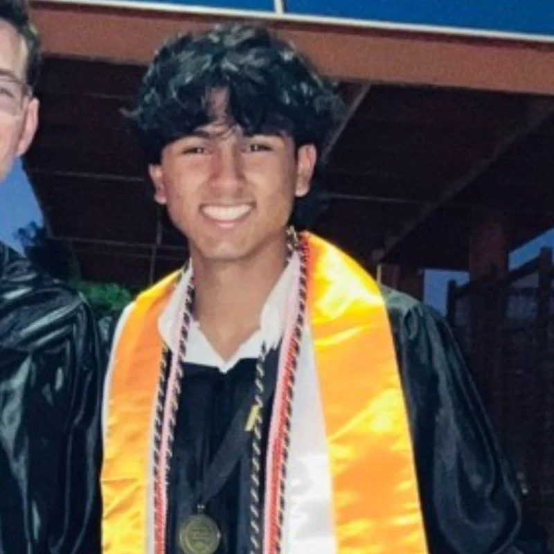 Young man wearing graduation gown and cap, smiling, with a gold sash and medal around his neck, standing outdoors during the evening.
