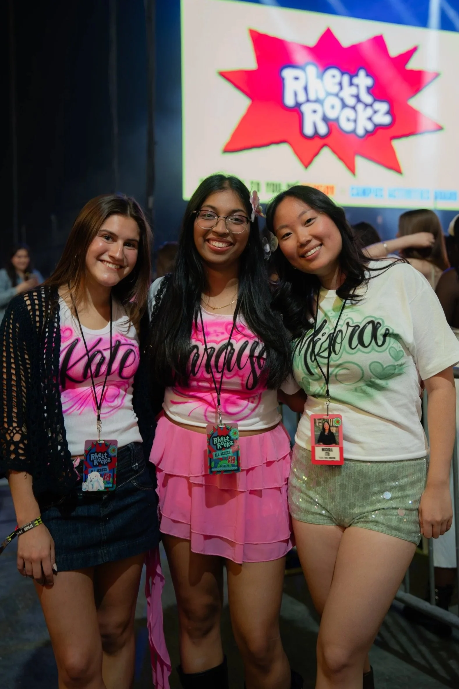 Three young women standing together at an event, smiling at the camera. They are wearing colorful casual outfits and event badges around their necks. A large sign behind them reads 'Rhett & Rockz'.