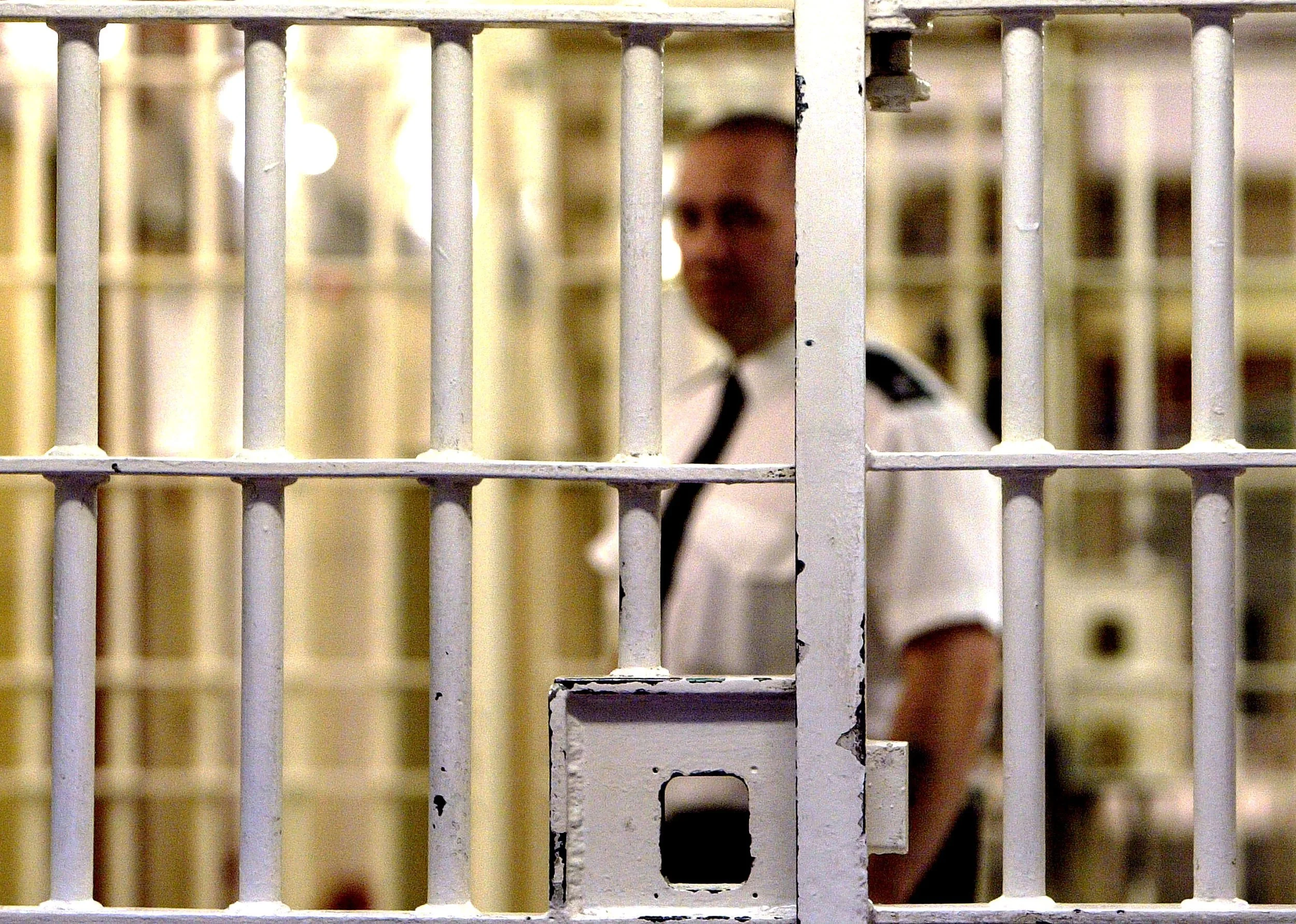 Prison cell with white metal bars, showing a blurred male inmate in a security uniform behind the bars.