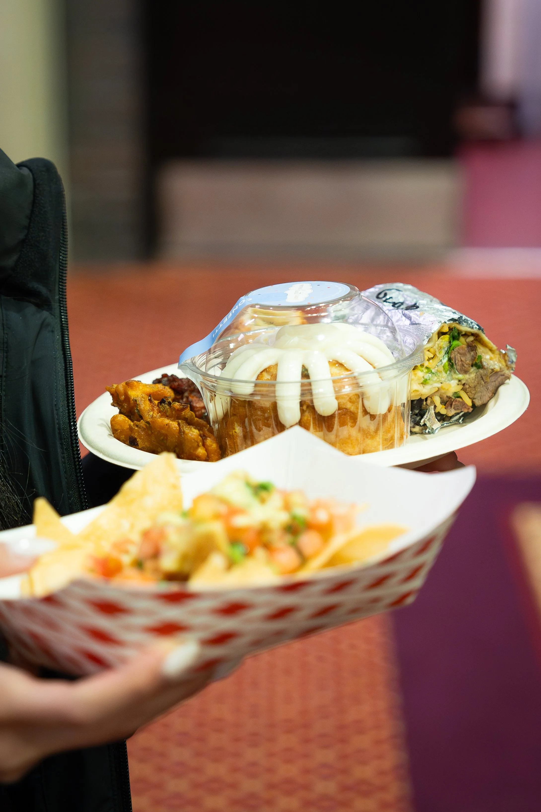 A hand holding a paper tray with various foods, including a cup with some food topped with white sauce, a wrap with meat and vegetables, and a side dish of seasoned vegetables, in an indoor setting.