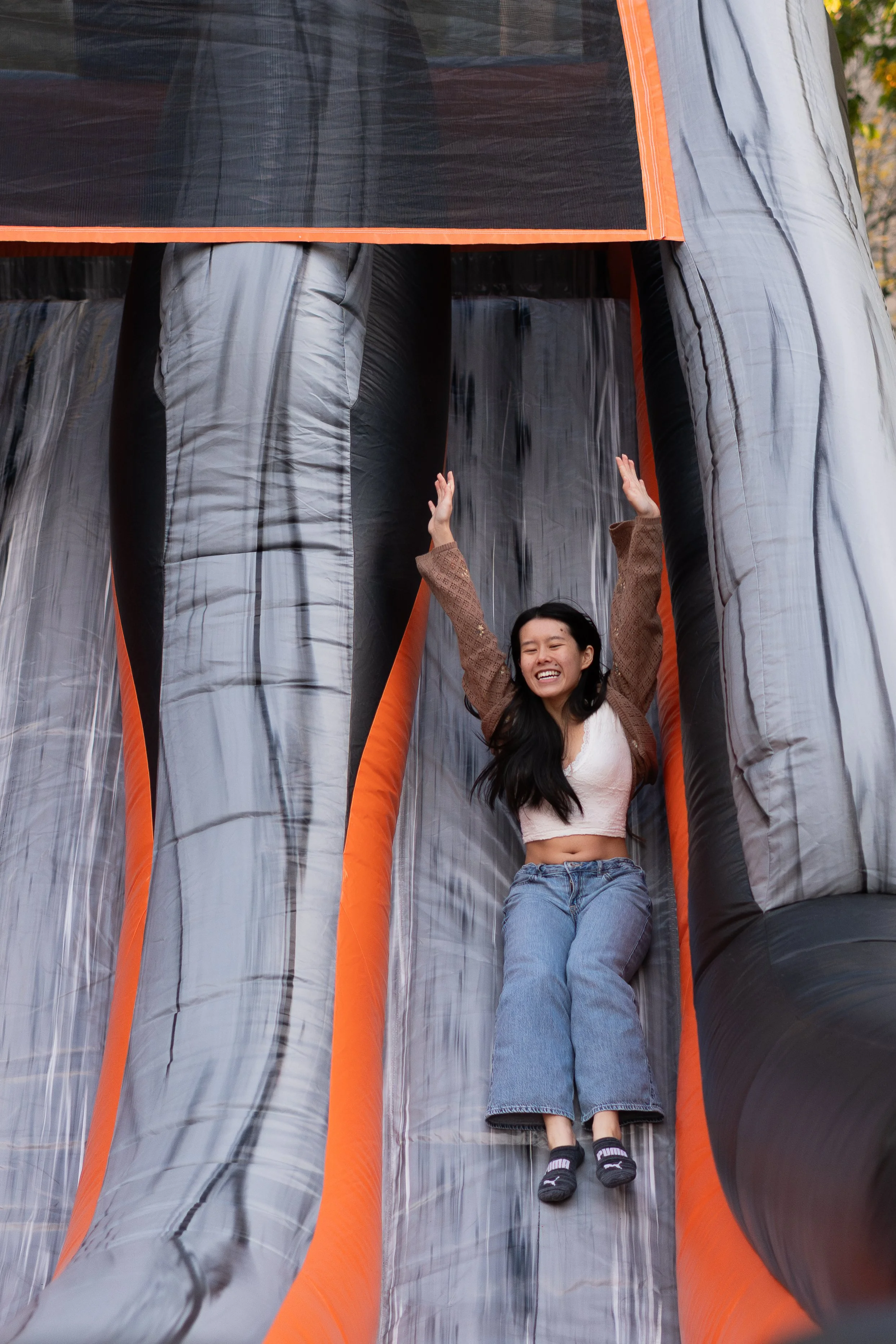 A woman with dark hair smiling and wearing a beige crop top and blue jeans, sliding down an inflatable water slide with her arms raised.