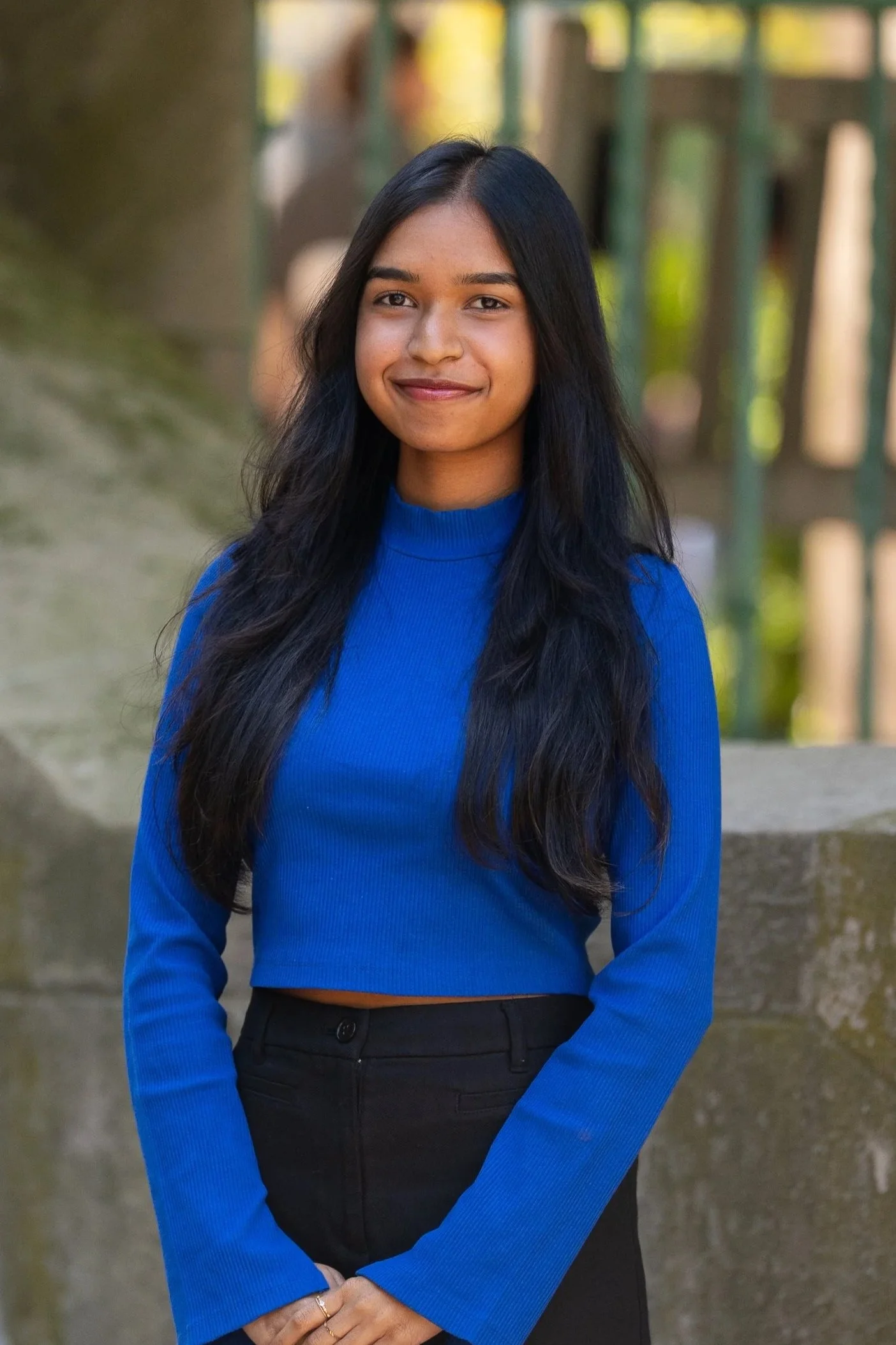 A young woman with long black hair, wearing a blue long-sleeve top and black pants, standing outdoors with a blurred background of greenery and a fence.