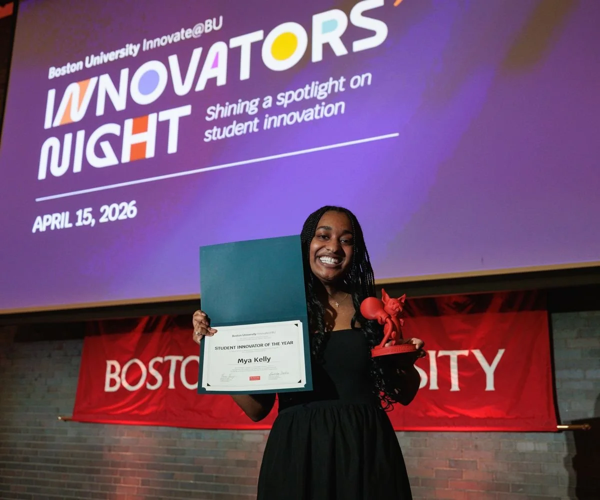 A young woman smiling and holding an award and certificate at Boston University Innovators Night event on April 15, 2026. The certificate reads 'Student Innovator of the Year' for Mya Kelly. In the background, a large screen displays the event details and a red Boston University banner is hung on the wall.