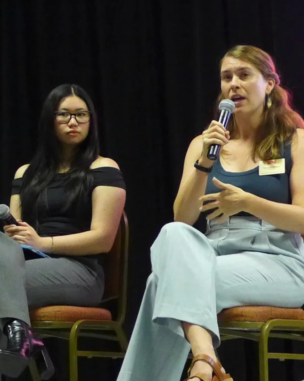 Two women sitting on stage. The woman on the right is speaking into a microphone, wearing a sleeveless top and light-colored pants. The woman on the left is listening, wearing glasses and a black off-the-shoulder top.