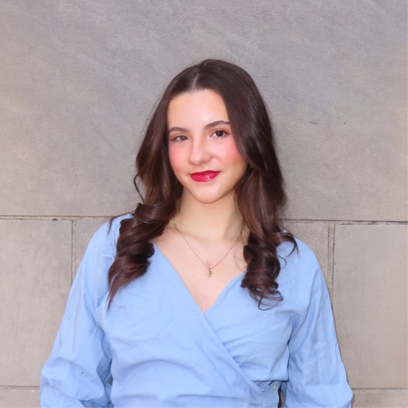Young woman with long wavy brown hair wearing a light blue blouse, standing against a gray stone wall.