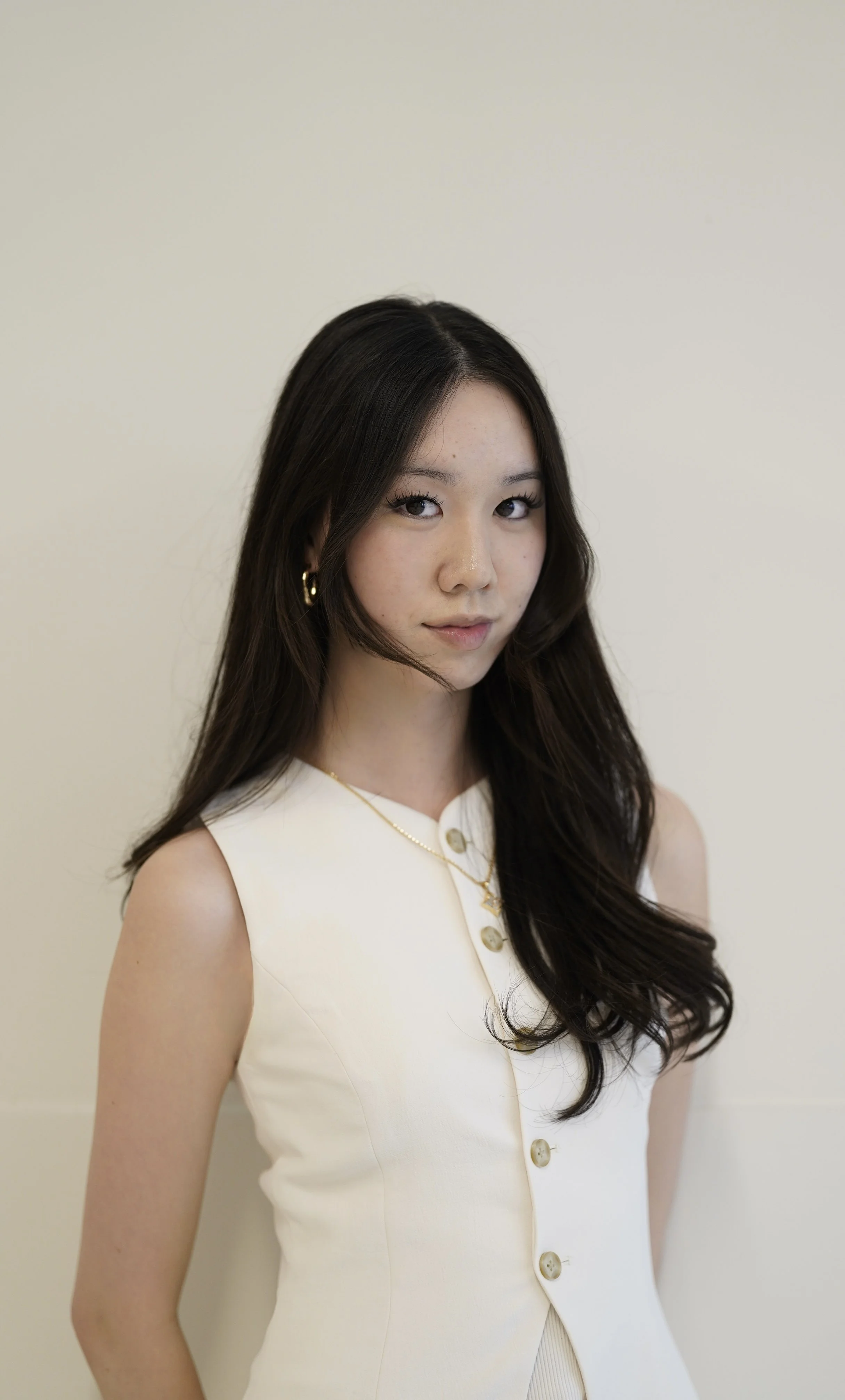 A young woman with long wavy dark hair, wearing a sleeveless white top with buttons and gold jewelry, standing against a plain light-colored background.