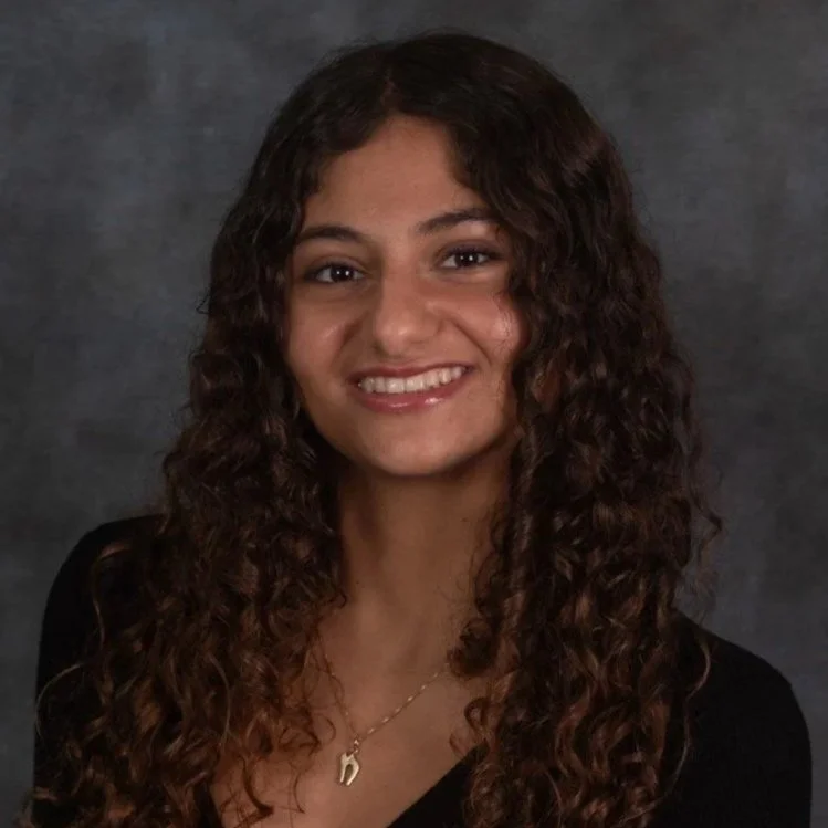 A young woman with curly brown hair smiling at the camera, wearing a black top and a gold necklace with a small pendant, against a dark gray background.