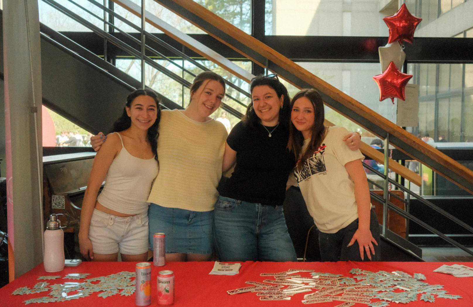 Four young women standing behind a red table with decorations, smiling and posing for a photo inside a building near the stairs, with balloons and outdoor visible through windows.