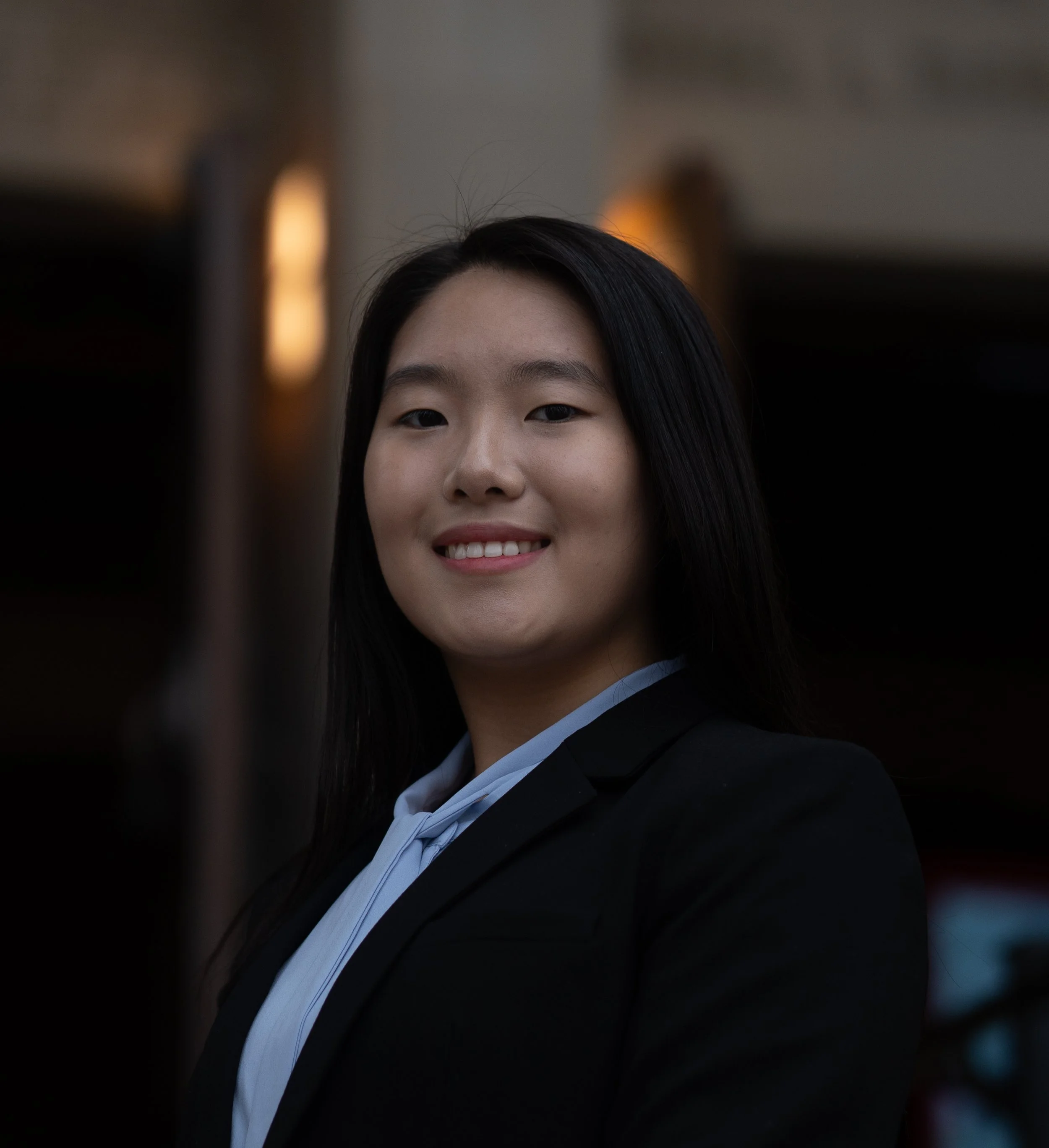 A young woman with long dark hair smiling in graduation attire, with a blurred background.