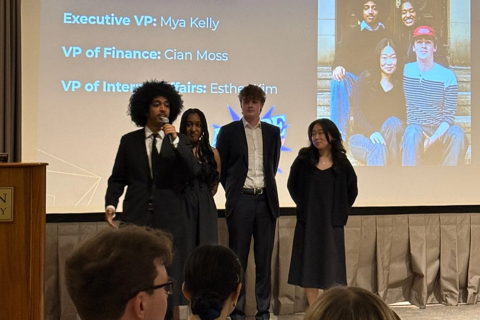 Four young professionals standing on a stage, with a presentation slide in the background listing their names and titles. The person on the far left is holding a microphone and speaking. Audience members are visible in the foreground.