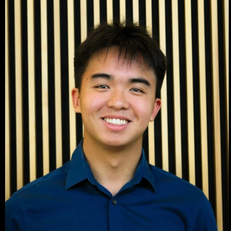A young man with short black hair, smiling, wearing a blue collared shirt, standing in front of a black and beige striped background.