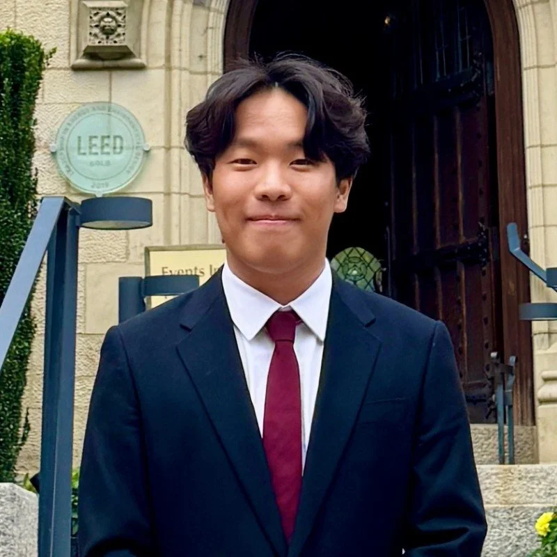 Young man in a dark suit and maroon tie standing outside a historic building with a large wooden door, stone steps, and greenery.