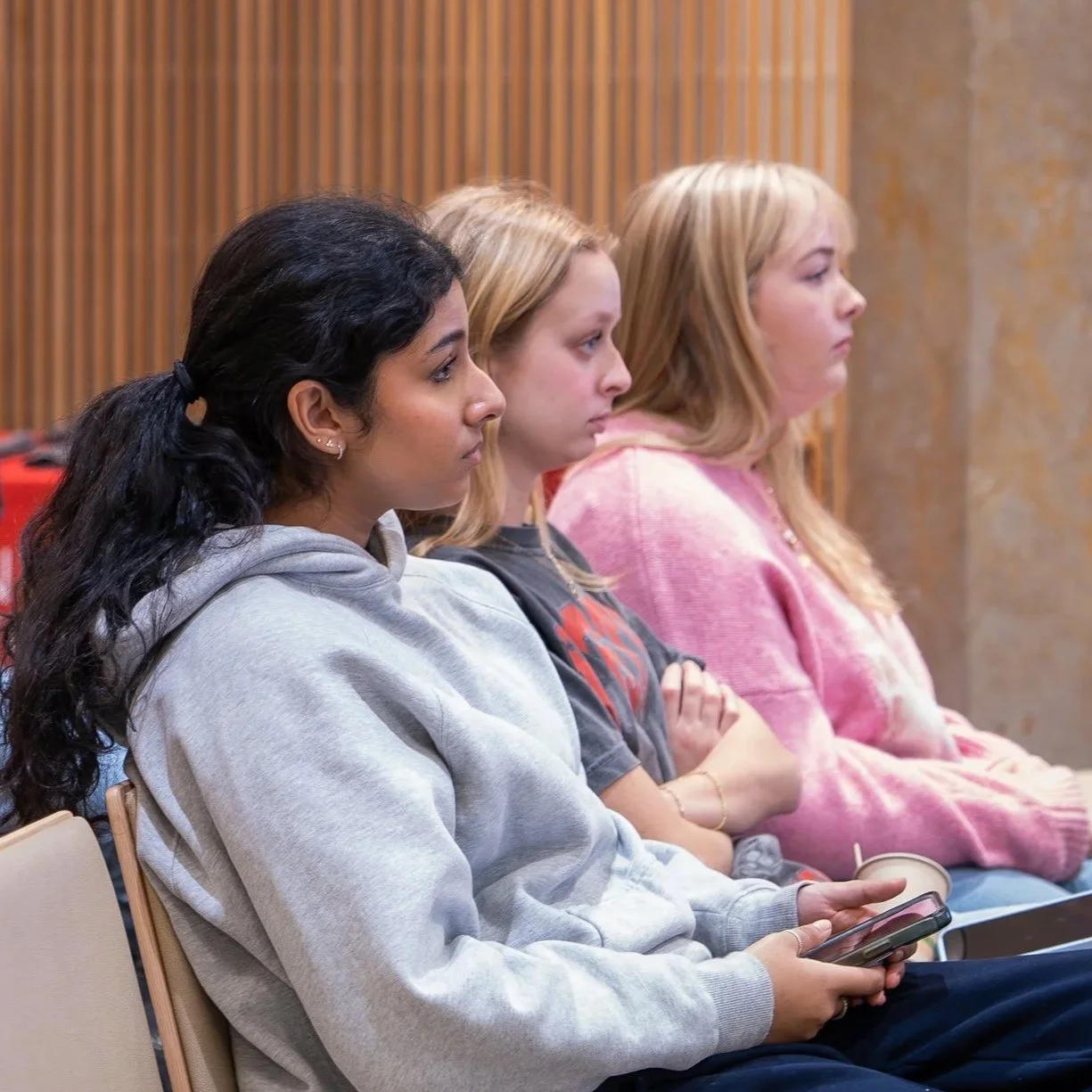 Three women sitting in a row, listening attentively, one holding a smartphone.