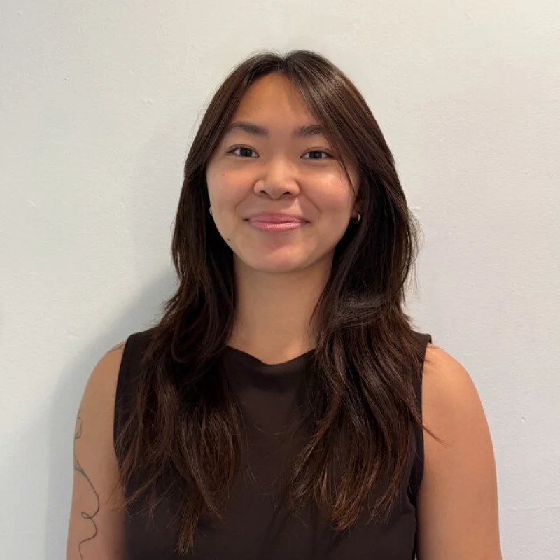 A young Asian woman with long brown hair, wearing a black sleeveless top, standing against a plain white wall, smiling gently at the camera.