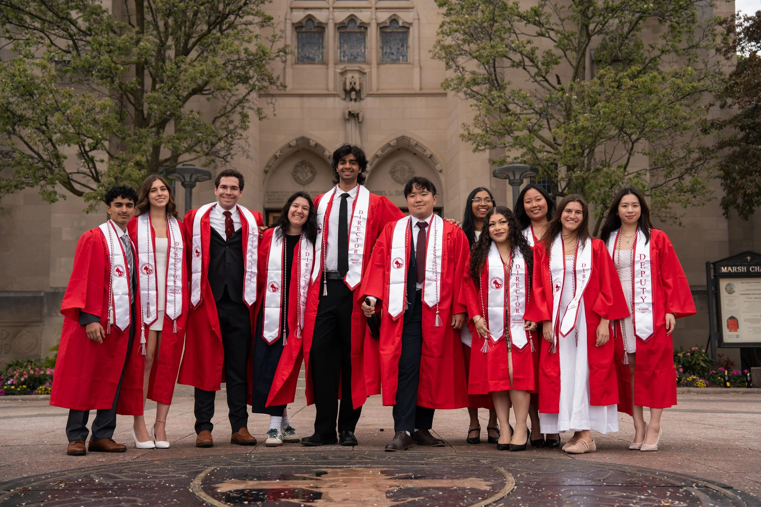 A group of twelve diverse students wearing red and white graduation gowns and sashes, standing outside in front of a historic stone building with large trees. They are celebrating graduation.