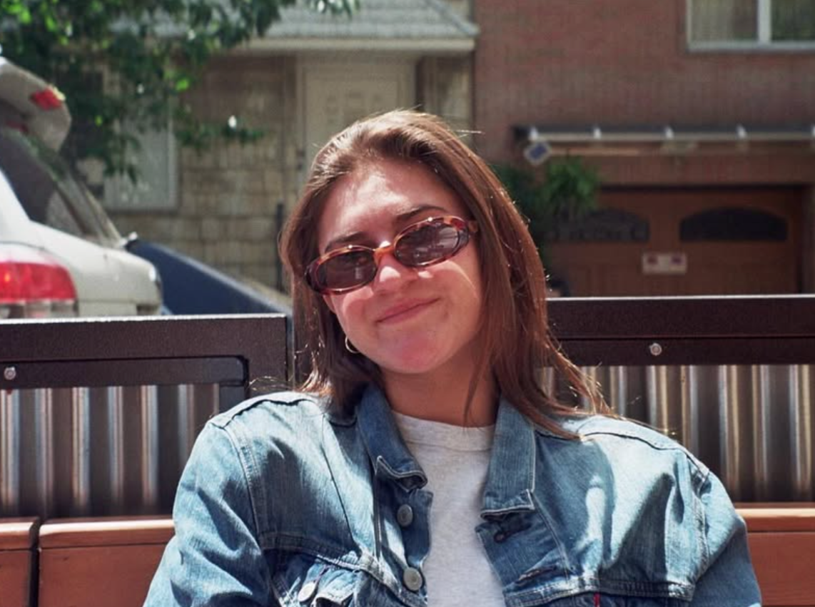 A young woman with brown hair, wearing sunglasses, a denim jacket, and a white shirt, smiling outside on a sunny day, sitting on a bench.