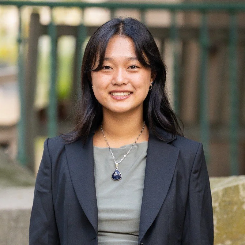 A young woman with shoulder-length black hair and earrings, wearing a gray shirt and black blazer, standing outdoors with a blurred background of a bridge and greenery, smiling at the camera.