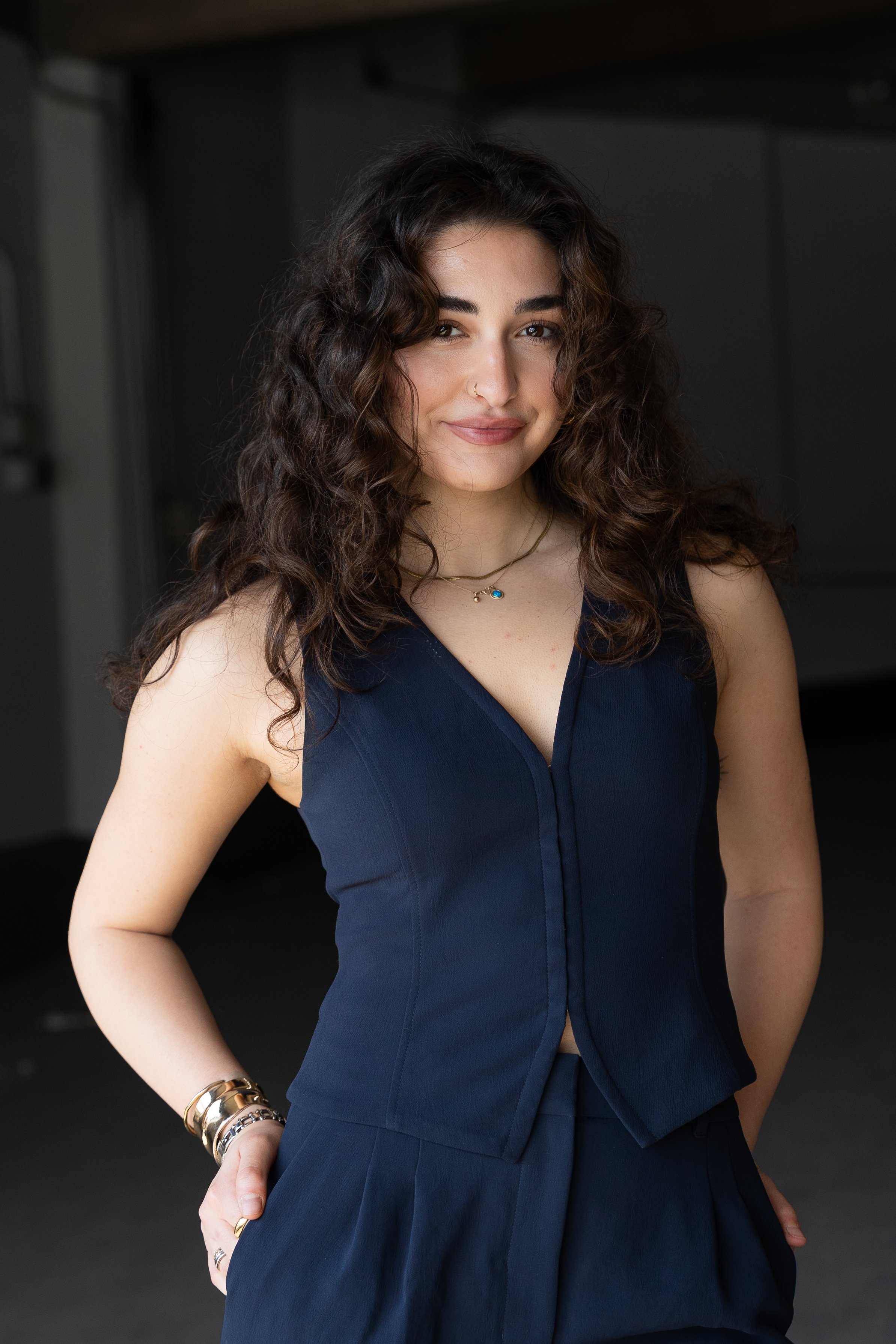 A woman with dark, curly hair wearing a navy blue sleeveless top, multiple bracelets on her left wrist, a simple necklace, and rings, smiling softly while standing against a dark background.