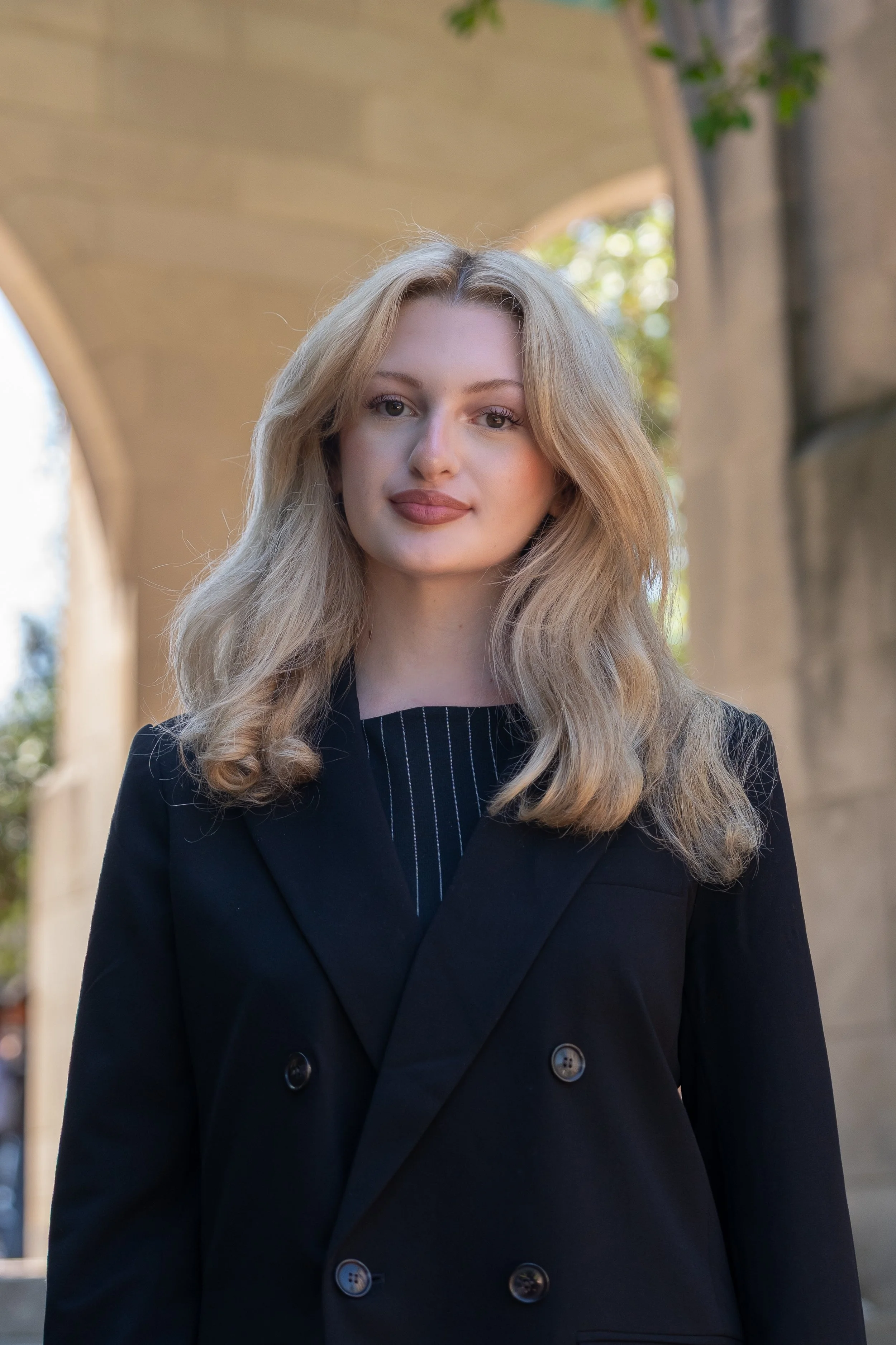 A young woman with long, curly blonde hair, wearing a black blazer over a white top, standing against a plain white wall.