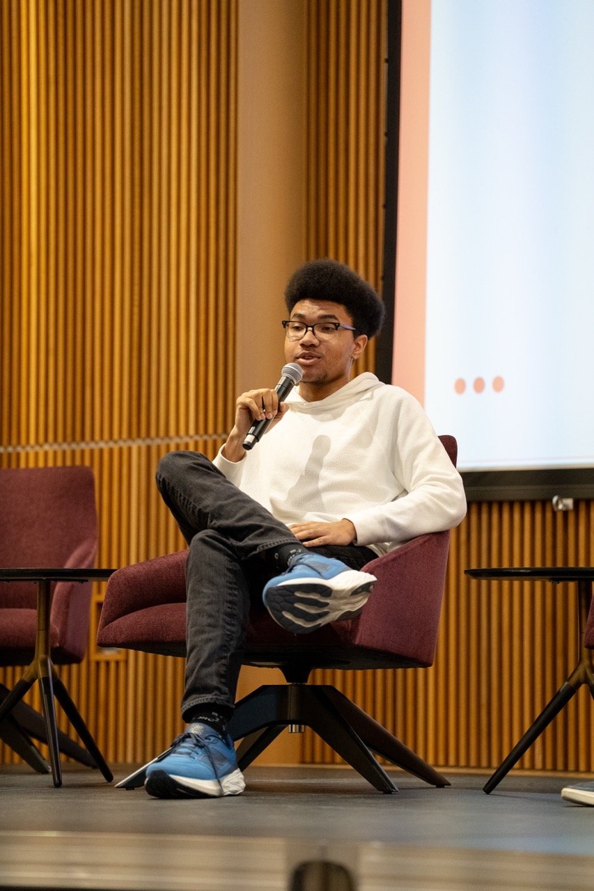 A young man with glasses and a tall hairstyle holding a microphone, sitting on a maroon chair with one leg crossed over the other, in a room with wooden wall panels and a white presentation screen.