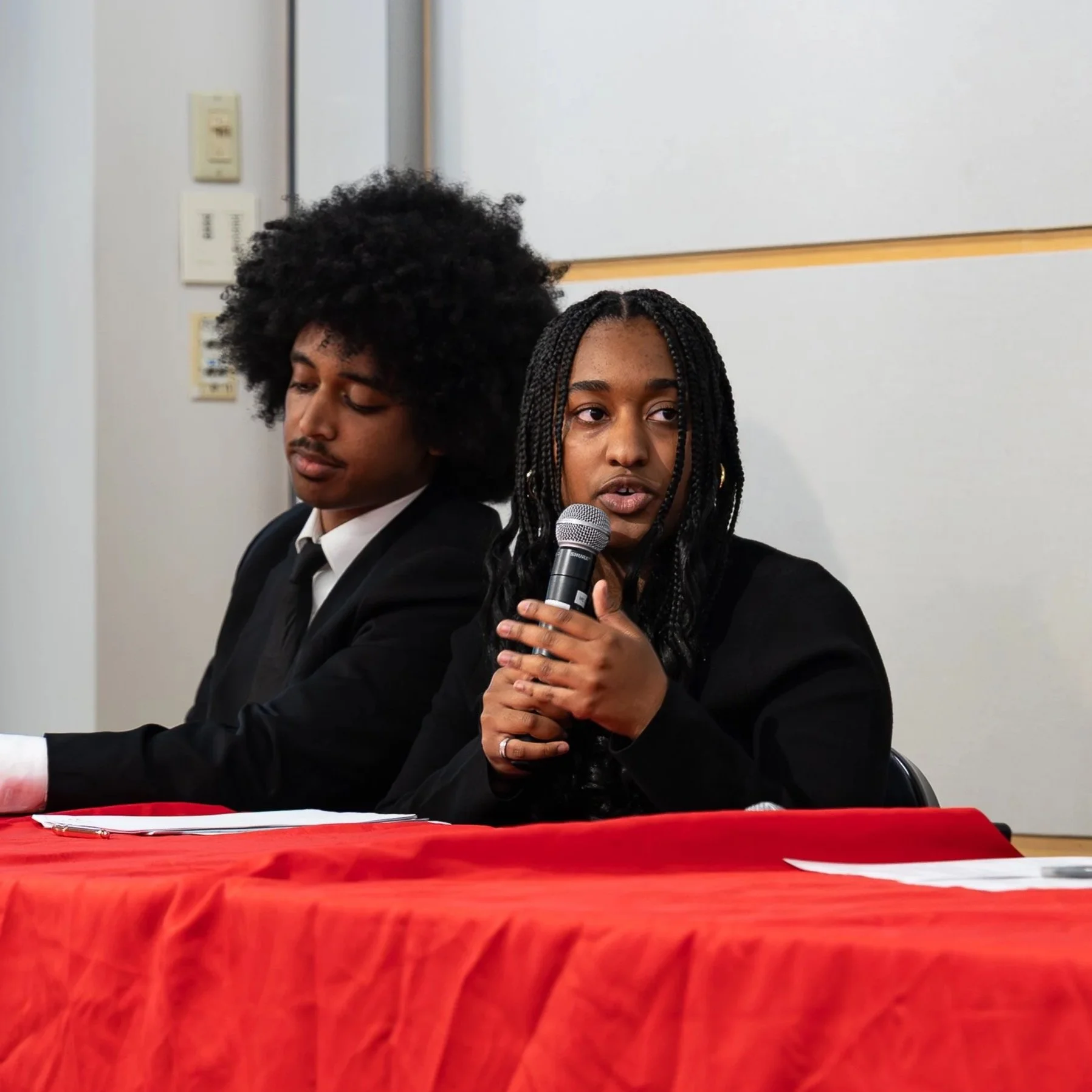 Two individuals sitting at a table with a red tablecloth in a room, one woman is speaking into a microphone and the other man rests with eyes closed, behind white wall with light switches.