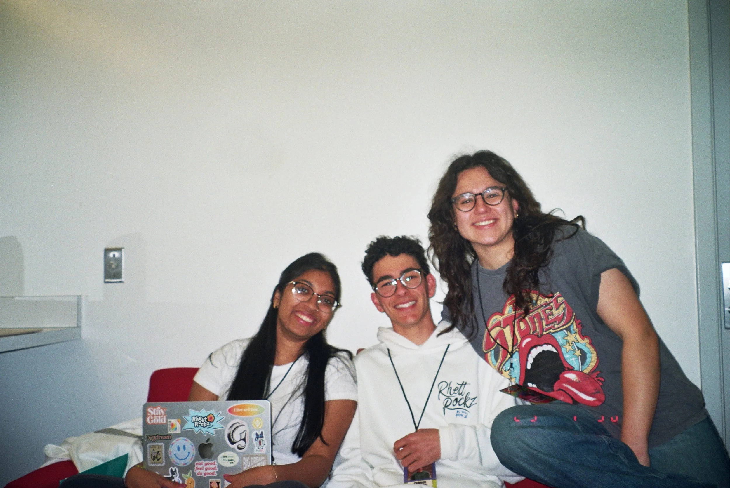 Four young people sitting and standing together in a room, smiling at the camera. The group includes three men and one woman, with a laptop covered in stickers in front of them and a plain wall in the background.