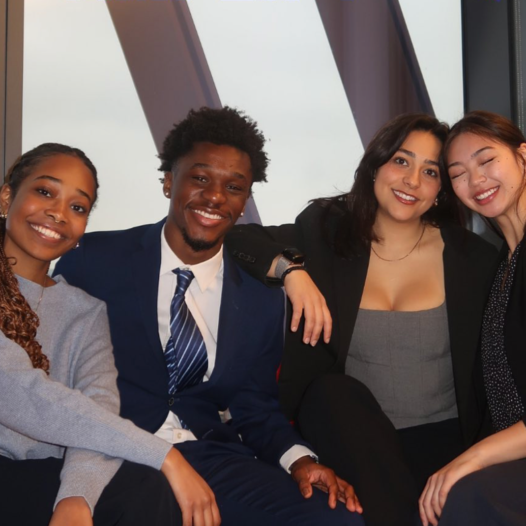 Four diverse young adults smiling and sitting close together, with one person having their arm around another, in an indoor setting.