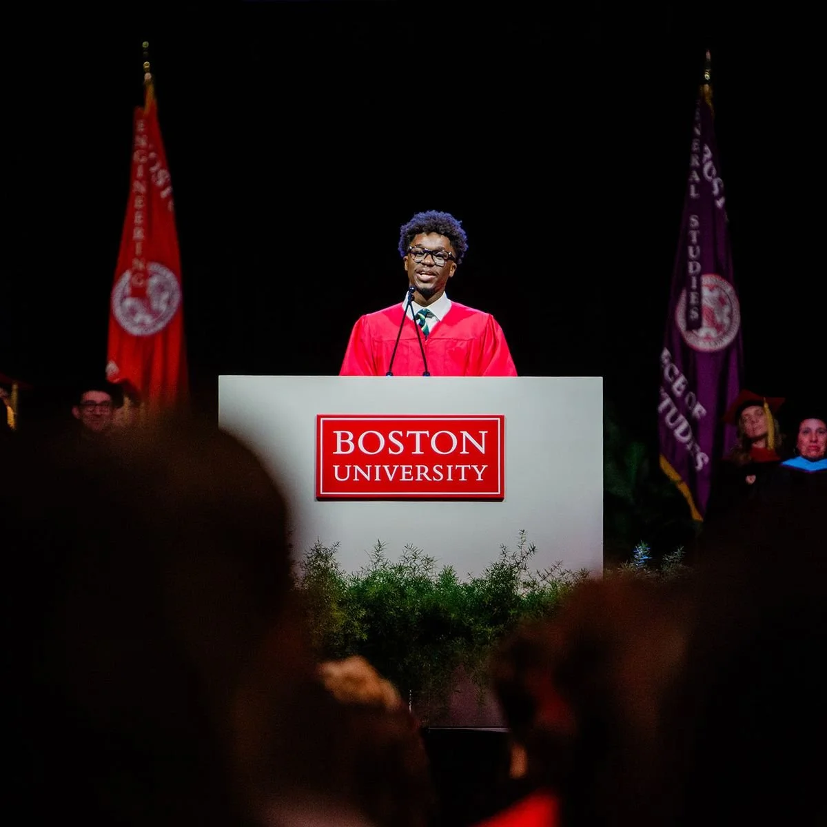 An African American male student in a red graduation gown giving a speech at a podium that says 'Boston University' during graduation ceremony. Flags and other graduates are visible in the background.