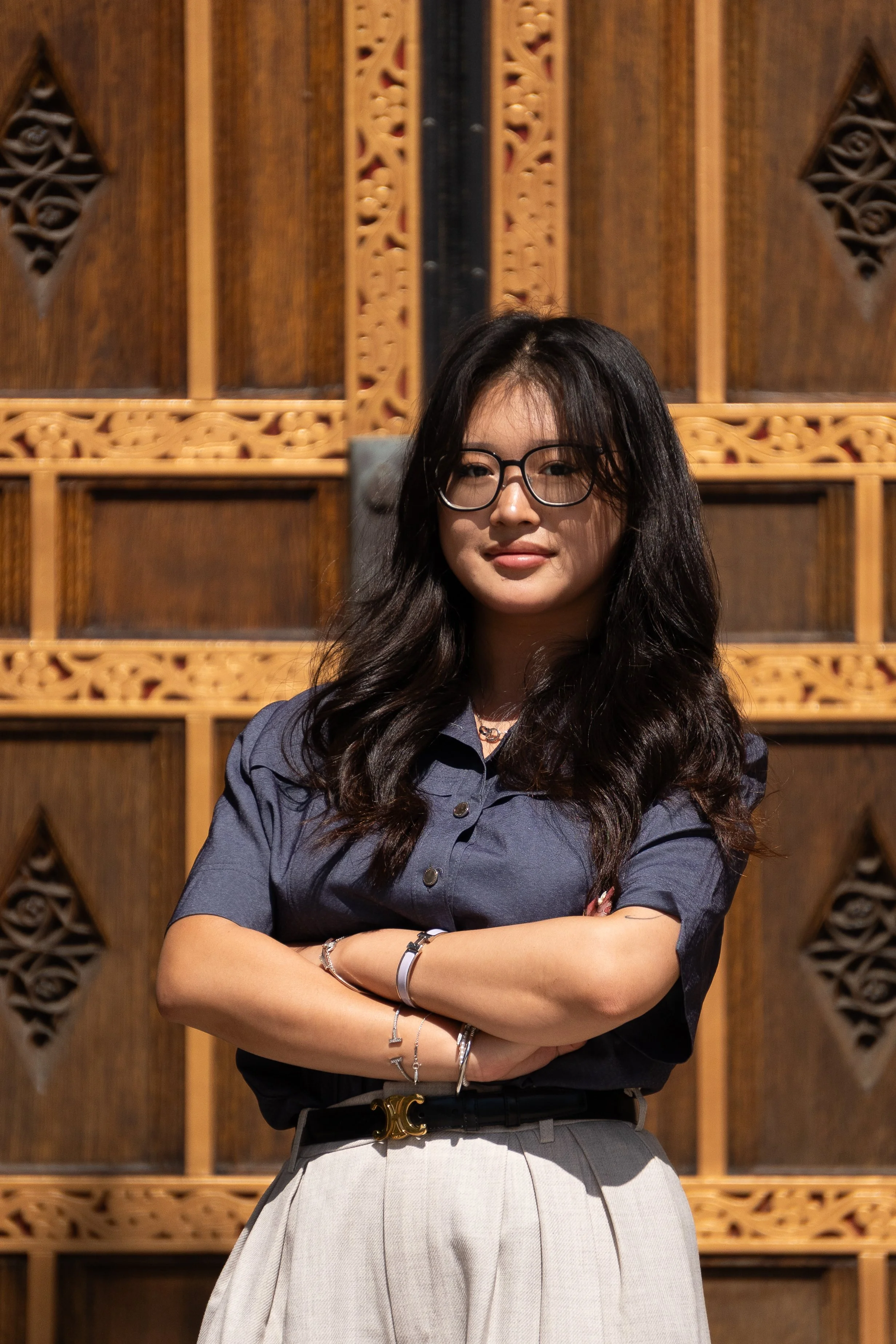 A woman with glasses and long dark hair, dressed in a dark gray shirt and light gray skirt, standing with arms crossed against a wooden background with intricate carvings.