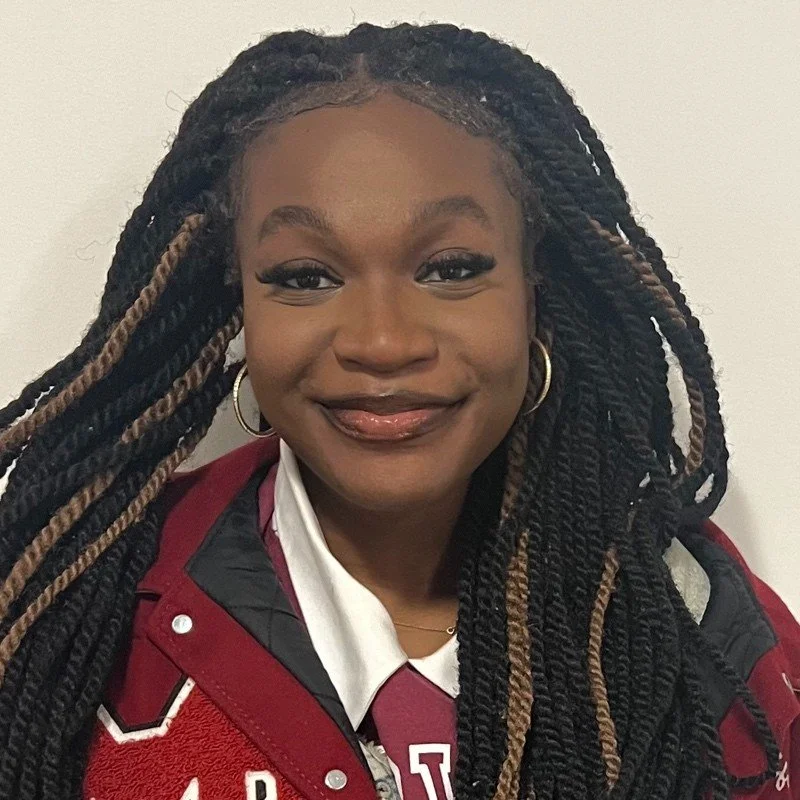 Portrait of a smiling young woman with long twisted hair, gold hoop earrings, wearing a maroon and black jacket over a white shirt.