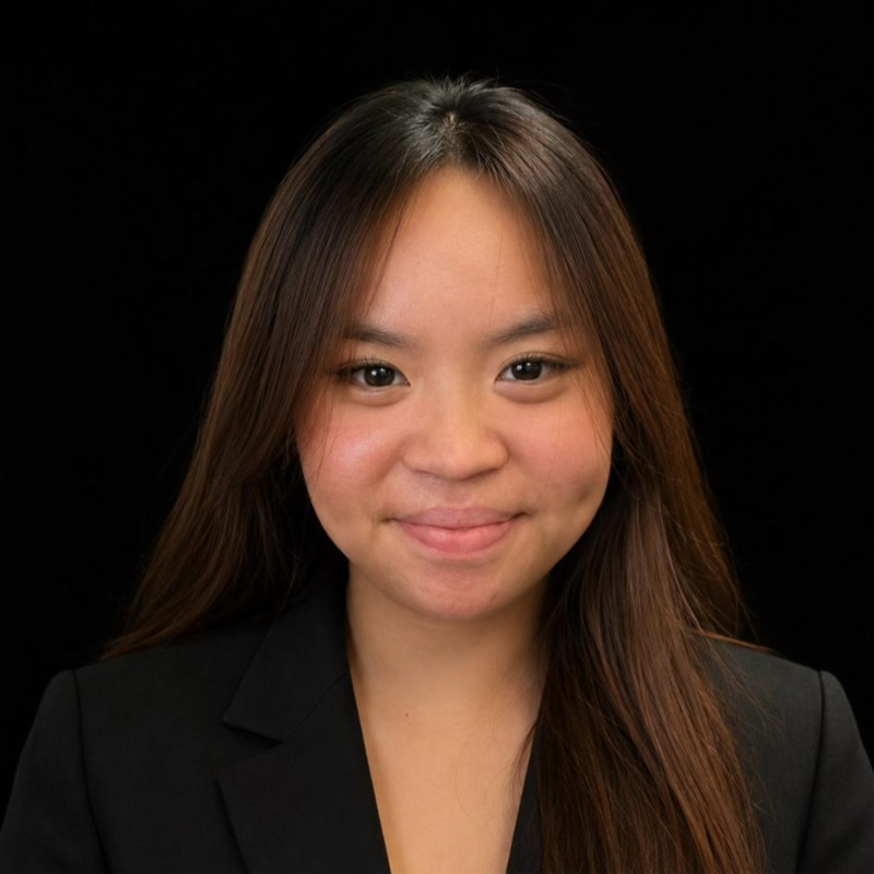 A young Asian woman with long brown hair in a black blazer, smiling softly against a black background.