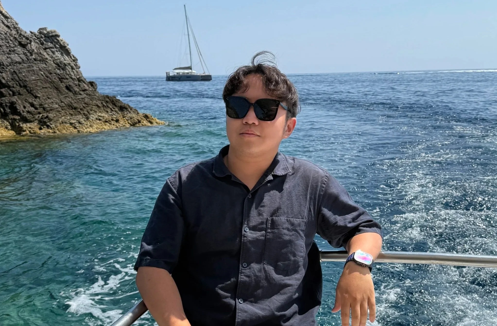 A young man wearing sunglasses and a gray shirt is sitting on a boat, with water, rocky cliffs, and another boat in the background.