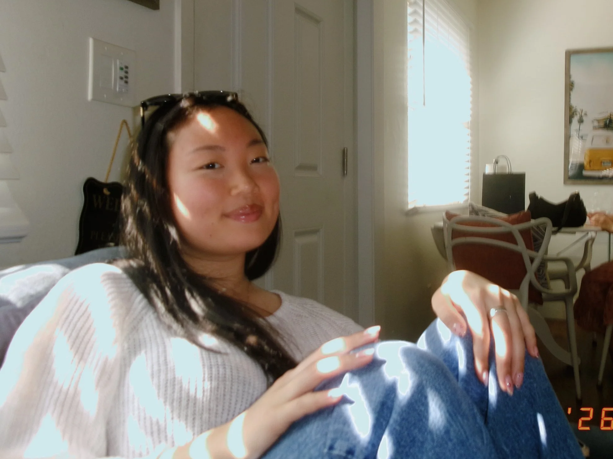 A woman with long dark hair, wearing a white top, smiling and holding a baby. The background shows a bright room with a window, blinds, a TV, and some chairs.