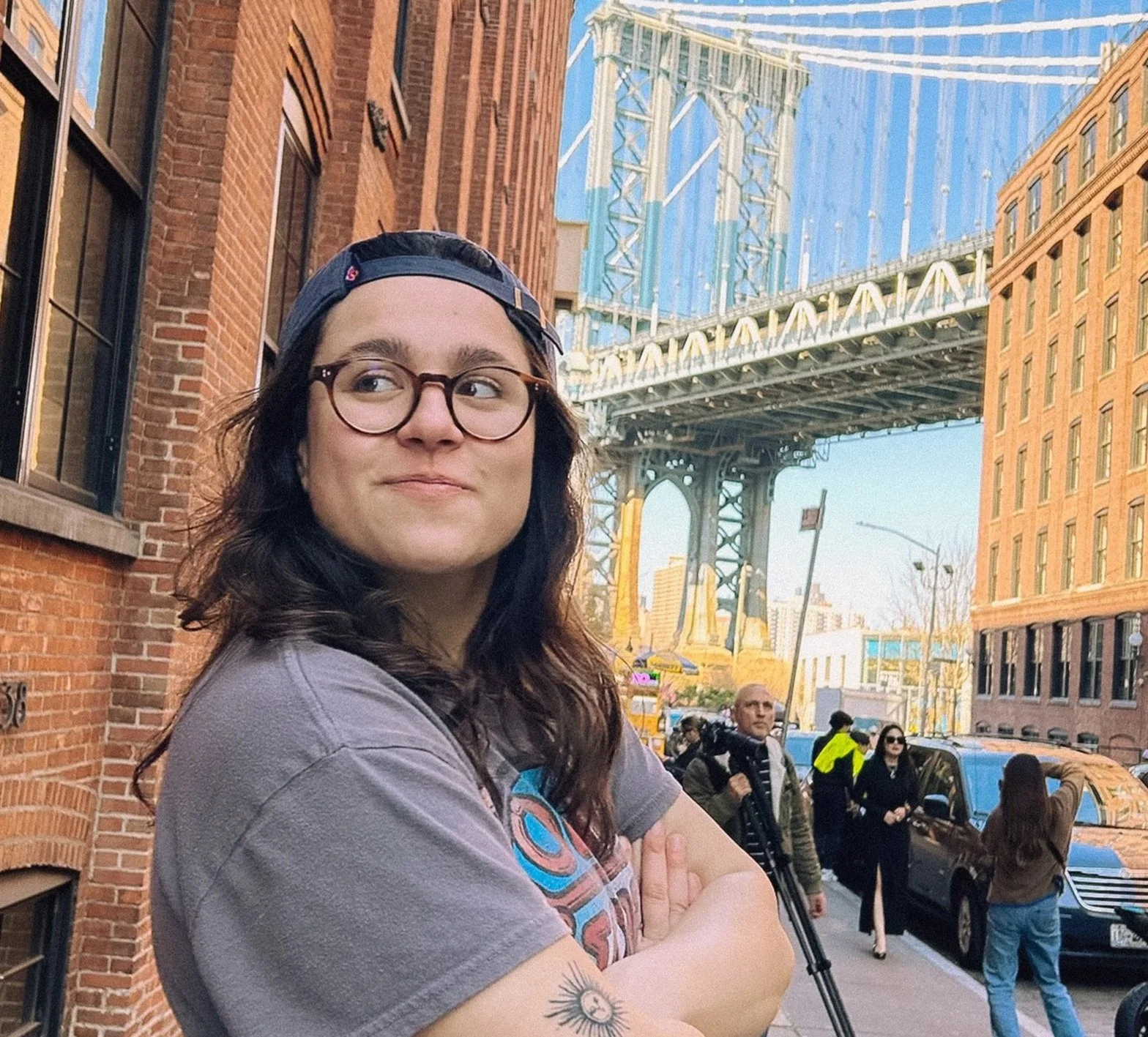 A woman with long dark hair, glasses, and a backwards cap standing on a city street with the Manhattan Bridge in the background. She is crossing her arms and looking to her left. Several people are visible on the street, including a man with a camera and a woman in black glasses.