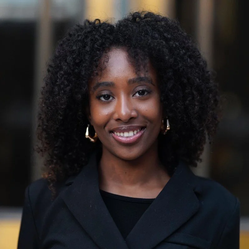 Close-up of a smiling Black woman with curly hair, wearing a black blazer and gold earrings, standing outdoors.