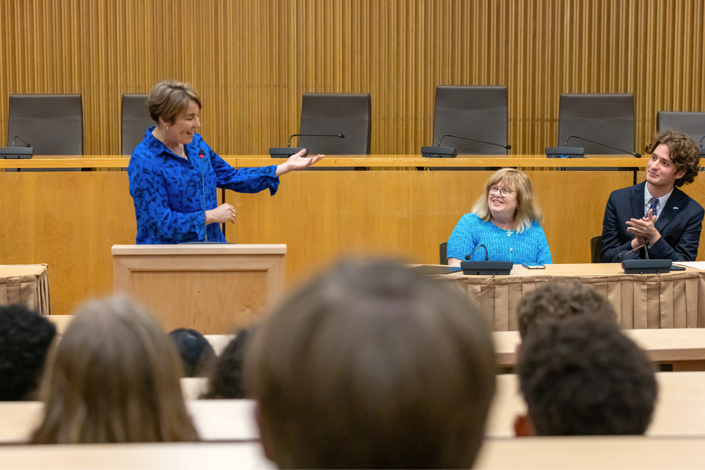 A woman in a blue patterned blouse is speaking at a podium in a room with wood-paneled walls. Two other people, a woman with glasses and a man with curly hair, are seated at a table, smiling and clapping in response. The image is taken from the back of an audience.