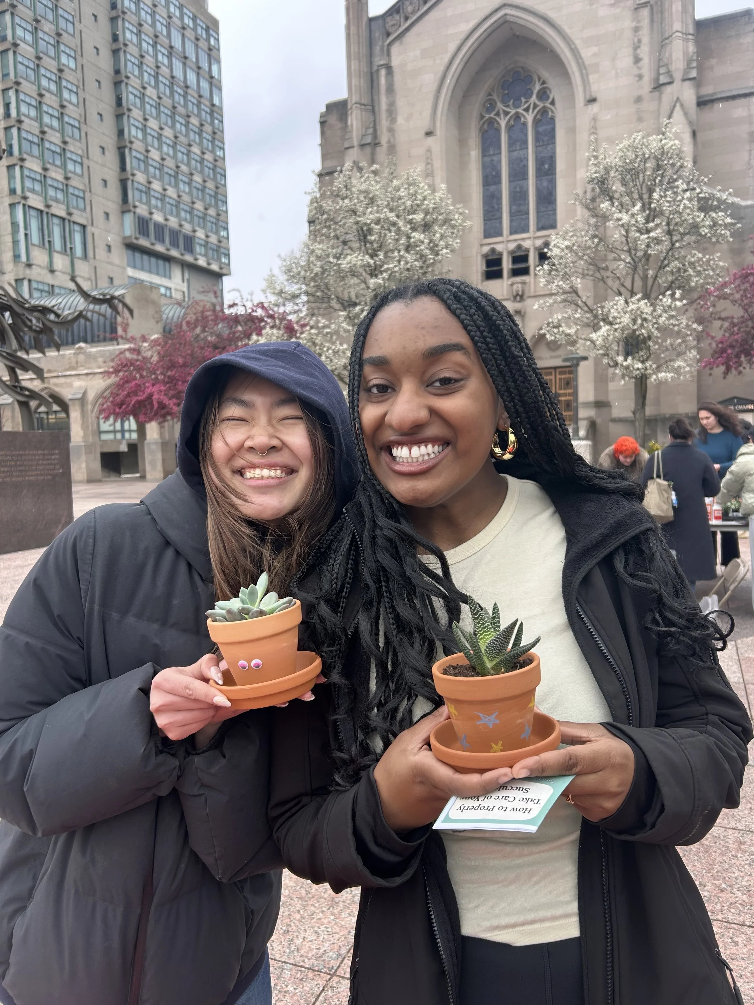 Two women smiling and holding small potted succulents with a historic building and trees with white and pink flowers in the background.