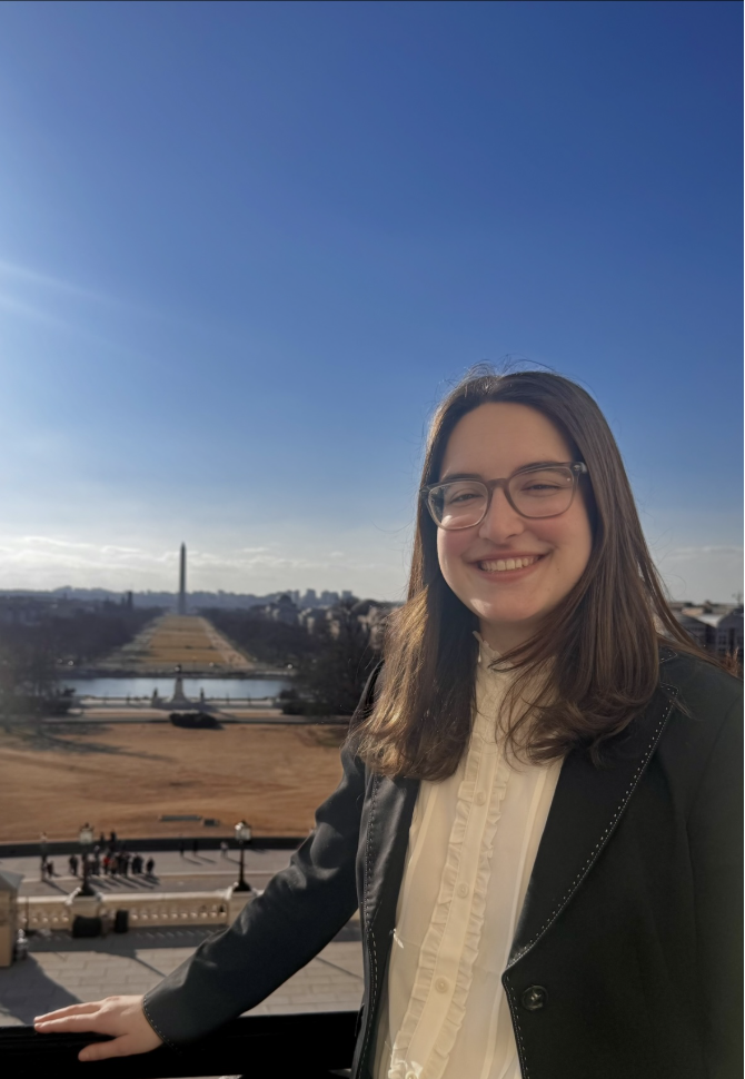 A young woman with glasses and long brown hair smiling outdoors with the Washington Monument and National Mall in the background on a sunny day.