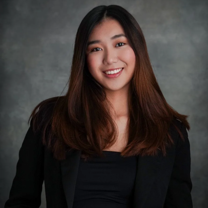 A young woman with long, straight brown hair wearing a black blazer and smiling against a gray textured background.
