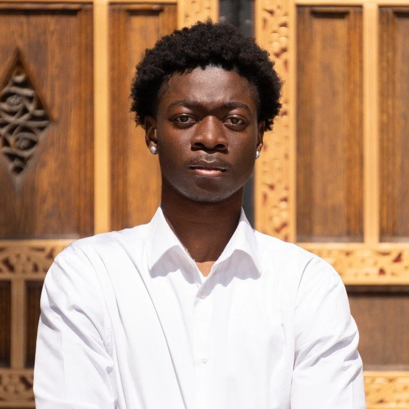 Portrait of a young person with dark skin, short curly black hair, wearing a white button-up shirt, standing in front of wooden decorative panels.