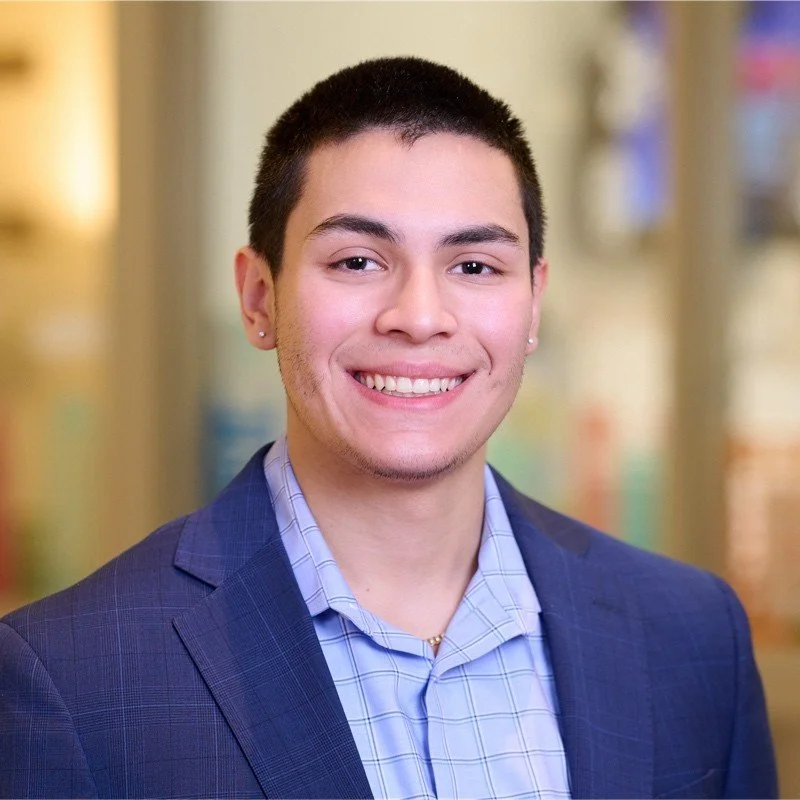 A young man smiling with short black hair, wearing a navy blazer and light blue checkered shirt, standing indoors with blurred background.