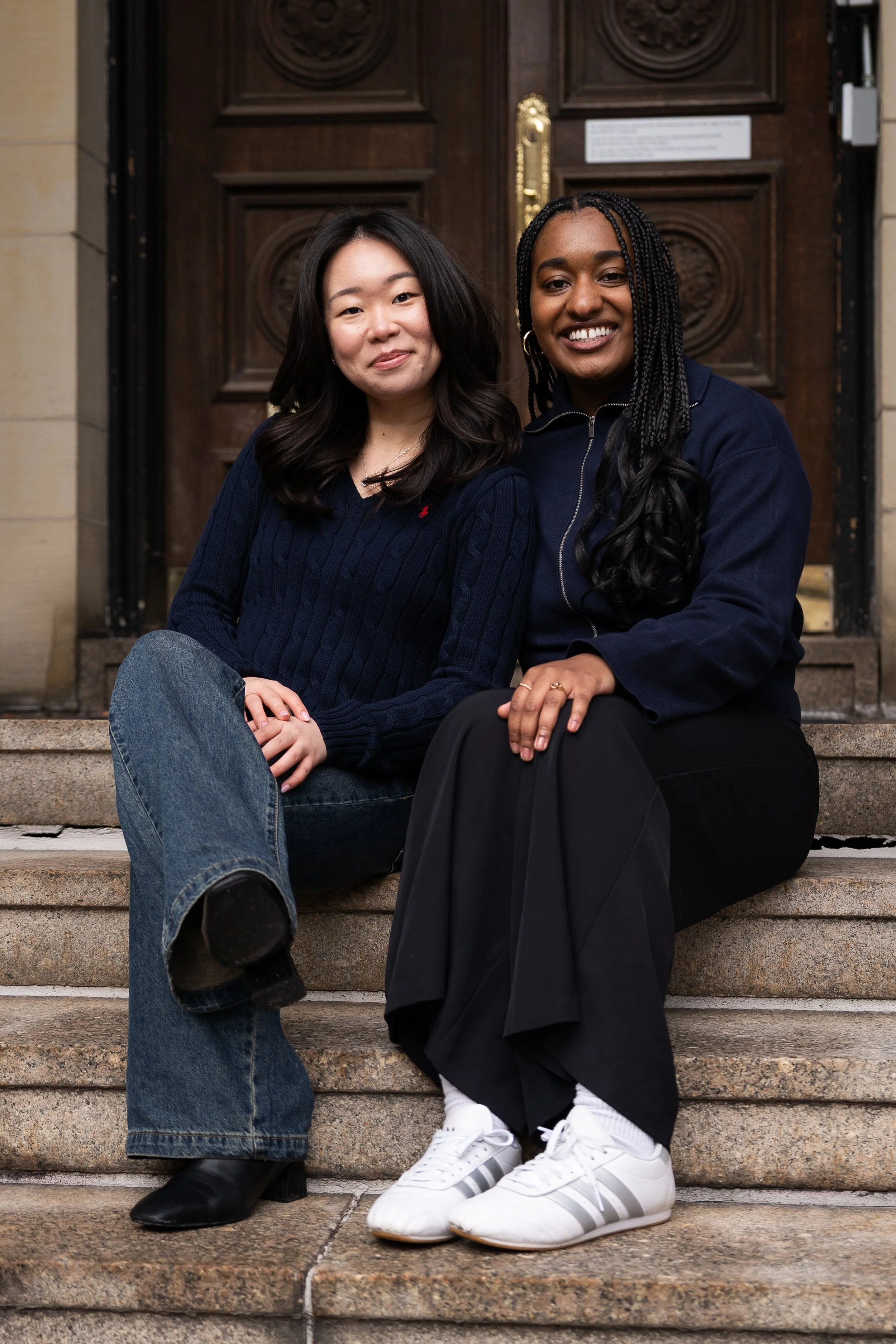 Two women sitting on steps outside a building with ornate wooden doors, smiling at the camera.