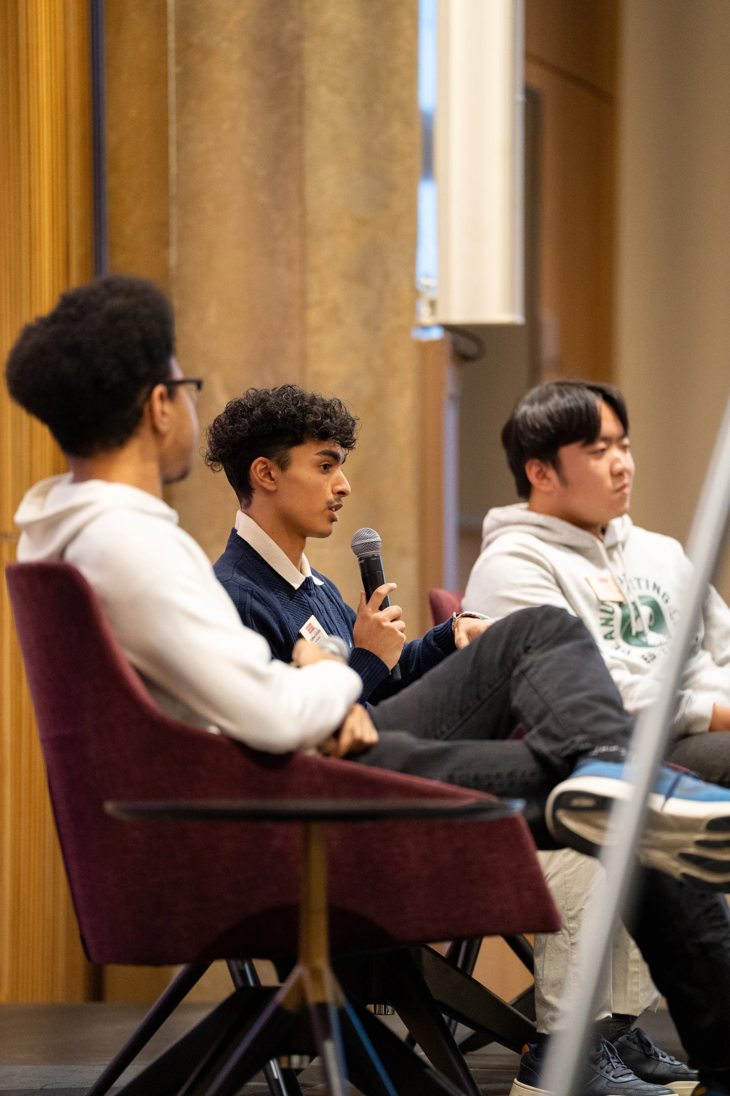 Three young men sitting in chairs during a panel discussion or presentation, one of them speaking into a microphone, in a conference room with wood-paneled walls.